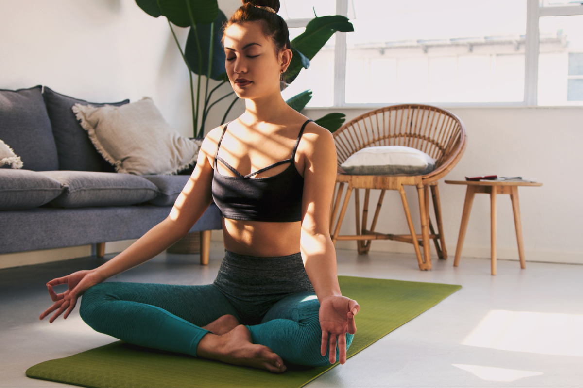 a young woman sits cross legged on a yoga mat with her eyes closed and hands in meditation pose