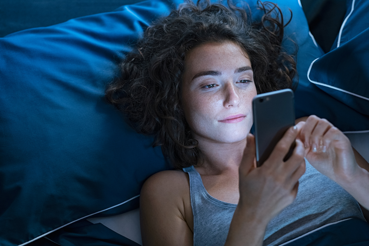 a woman lays in bed scrolling on her phone instead of sleeping