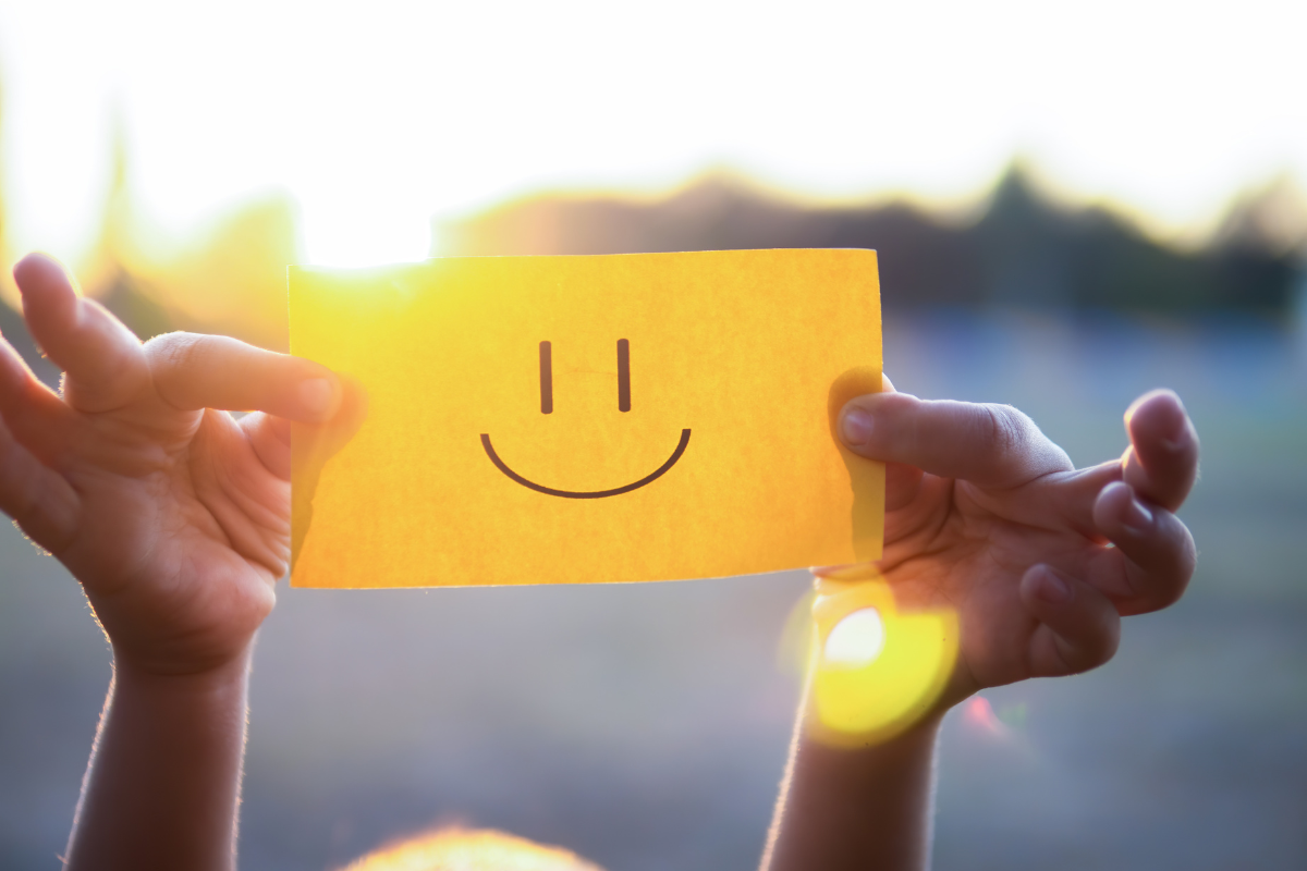 Pair of hands holding up a yellow slip of paper with a smiley face drawn on it (mouth and two eyes)