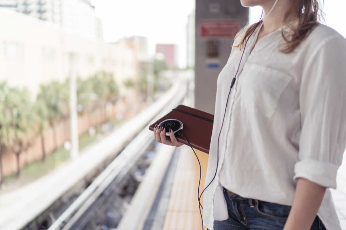 a woman stands in front of a commuter train, you cannot see her face but she is wearing xen headphones and carrying a journal and the xen device in her hand