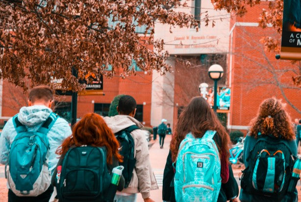 Five college aged young adults walk into a campus building that is red and white brick and surrounded by lovely trees that are orange and gold with fall leaves
