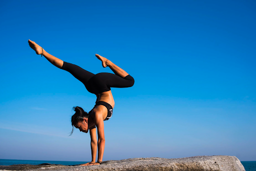 young woman doing a complex yoga pose on the top of a mountain cliff offset by a beautiful blue sky