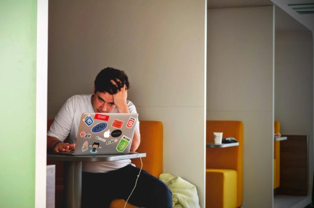 Young man sitting in colorful cubicle hunched over his laptop with a hand on his head and a visibly stressed look on his face. 
