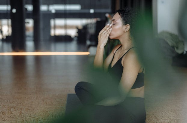 Woman sitting in a calm, cross-legged pose, on a yoga studio floor while covering her mouth and nose with her hand.