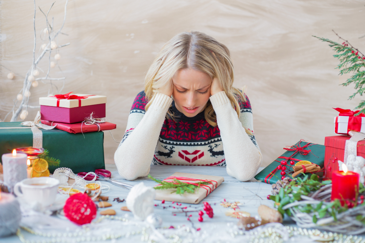 a woman sits defeated with her head in her hands in front of a table in disarray with holiday items like wrapping paper, baking items, and gifts