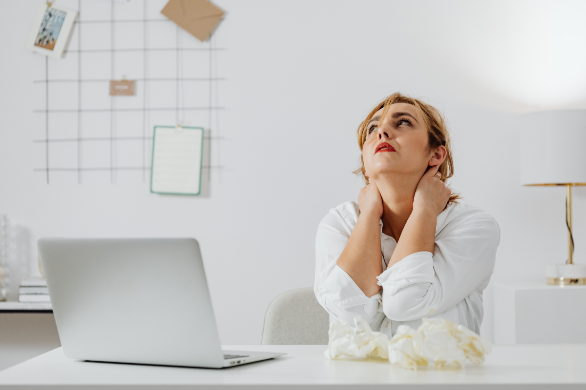 a woman sits at a desk with her hands on her head looking upward in a stressful position
