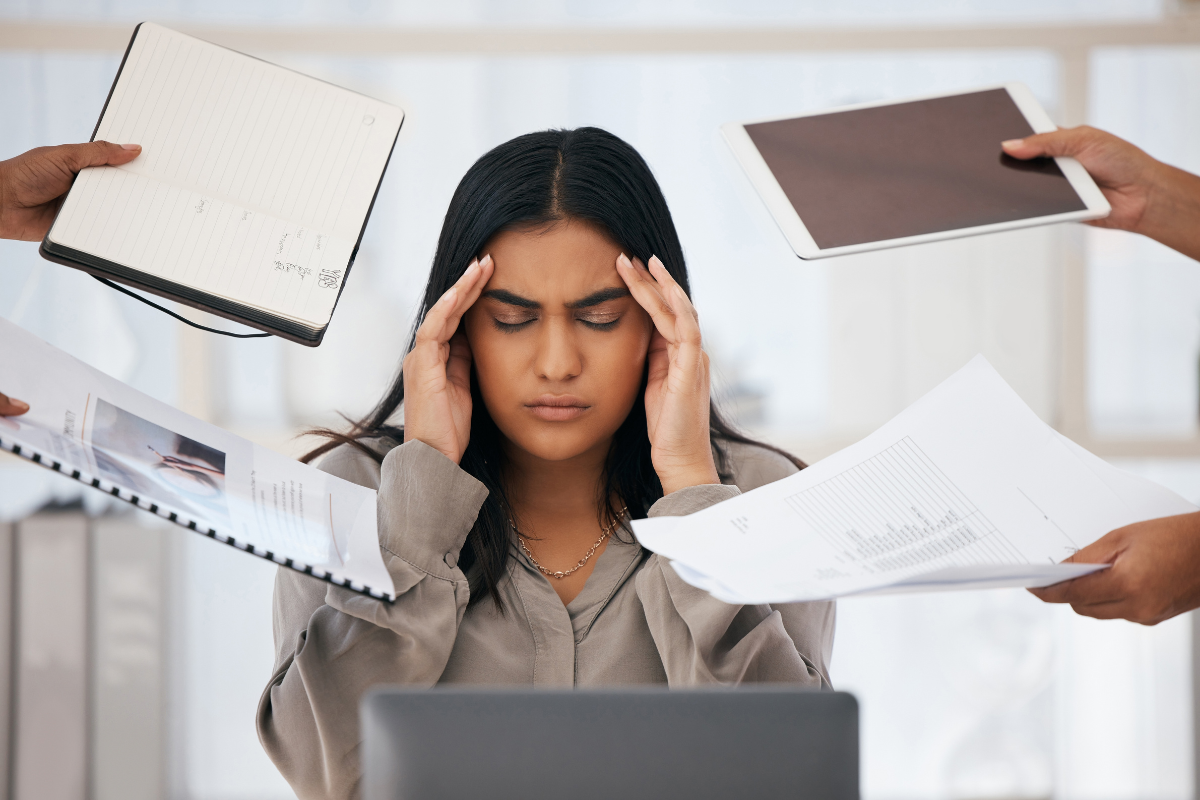 a stressed out woman at work sits at her laptop with hands on her head as other hands within the frame shove papers, binders, and ipad into her personal space
