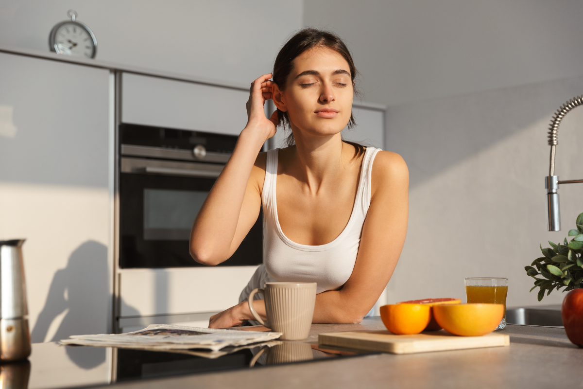 a woman stands sleepily at a kitchen counter, before her is a cup and some oranges sliced, her eyes are closed and natural sunlight illuminates the scene