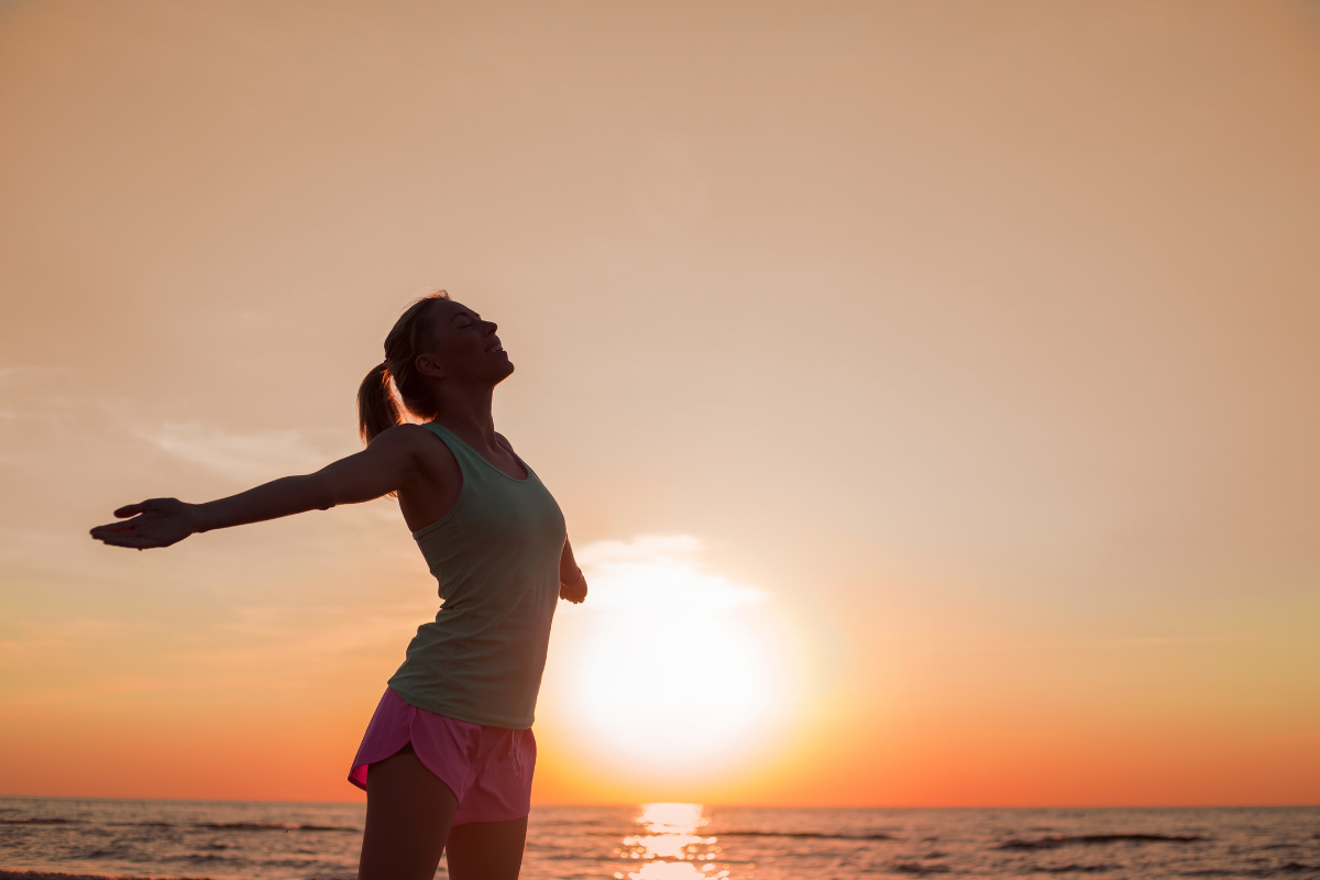 A fit looking woman stands, arms outreached, on a beach in front of a beautiful golden sunset