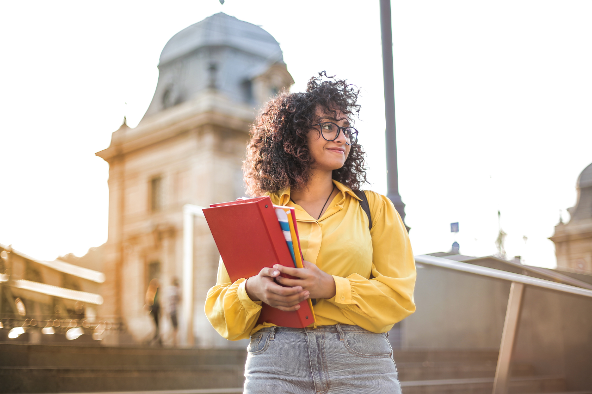 A young college aged woman stands on campus holding her books