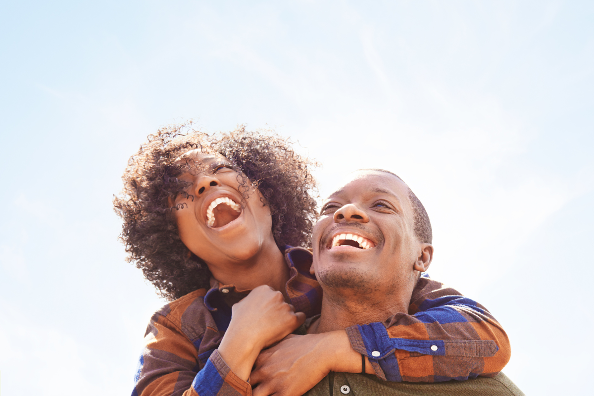 Photo of a woman with her arms wrapped around the shoulders of a man, they are both facing the camera and laughing gleefully at a subject unknown and off camera