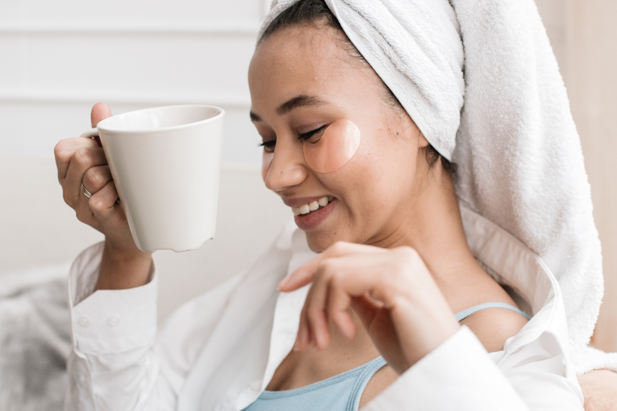 A woman with a happy expression on her face holds a cup of tea with a robe on and towel on her head she is relaxing and loving it