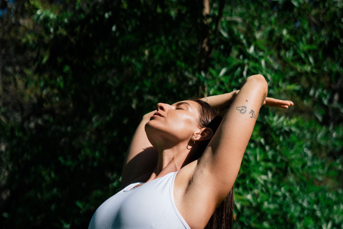 a woman offset by a lush green background of leaves stretches her arms over her head looking up toward the sky with a look of tranquility on her face