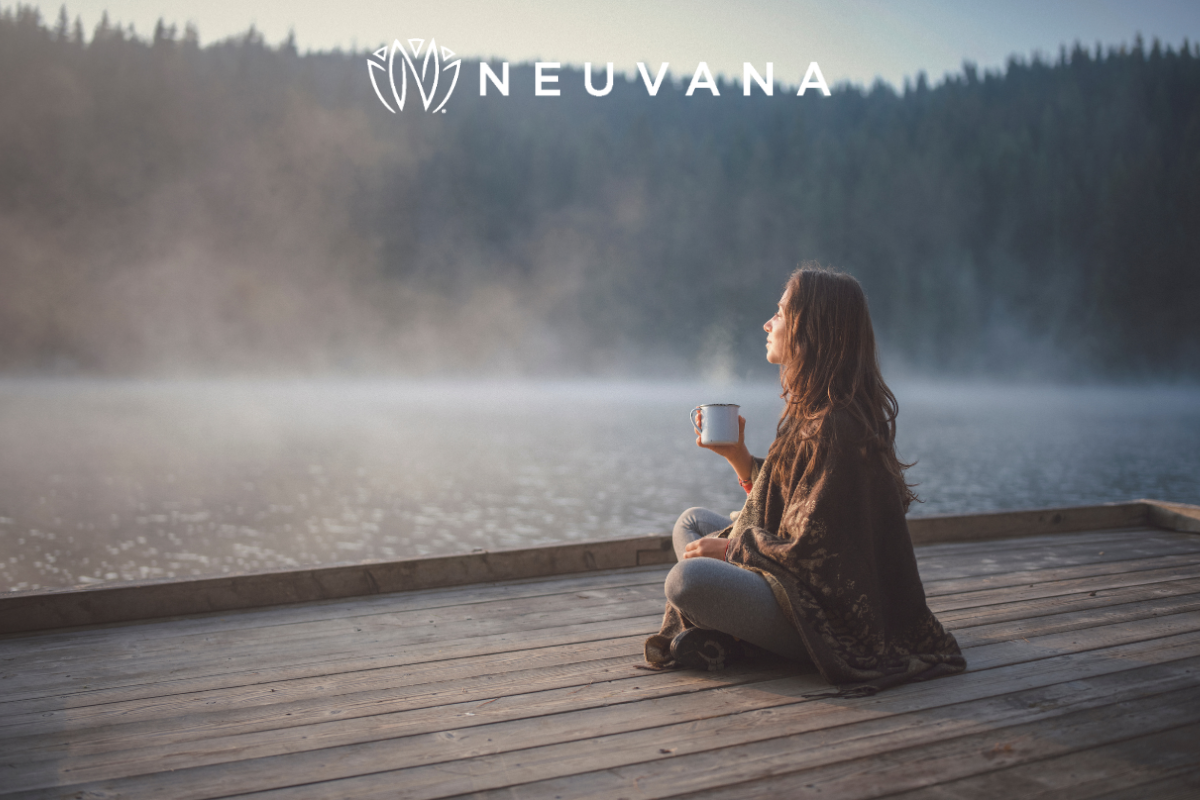 A woman sitting on a wooden dock by a lake, holding a cup of coffee, wrapped in a blanket, with a serene, misty forest in the background and the Neuvana logo above