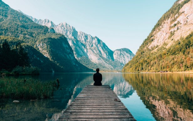 A lone person sits on a weather worn wooden dock on a reflective lake that is bordered  by majestic mountain ranges in various natural colors of steel gray, lush green, tans, oranges, and yellow the entire scene is surrounded by a crystal clear blue sky