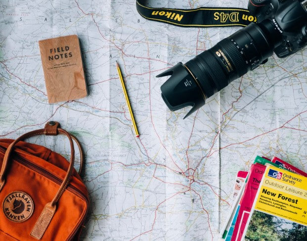 Travel essential items including a black nikon d4s camera with extended lens, brown field notes book, yellow #2 pencil, burnt orange kanken brand backpack, and assorted travel guidebooks in assorted colors of yellow, pink, and green lay on top of a map.